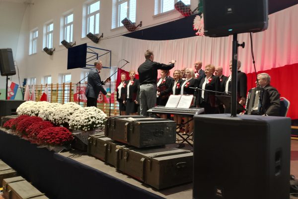 Zespół śpiewający na scenie z biało-czerwoną dekoracją i kwiatami podczas Festiwalu Pieśni Patriotycznych.