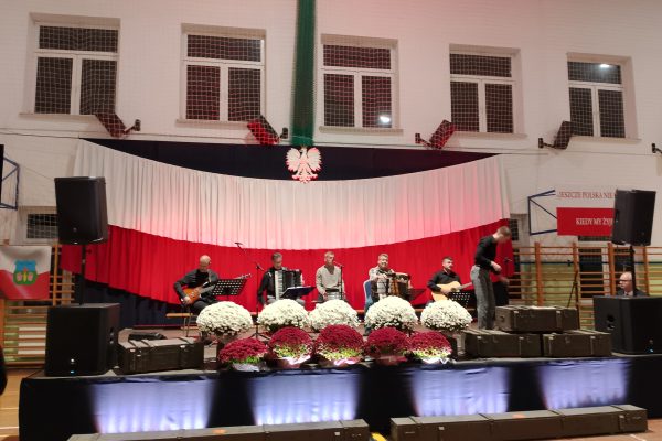 Zespół śpiewający na scenie z biało-czerwoną dekoracją i kwiatami podczas Festiwalu Pieśni Patriotycznych.