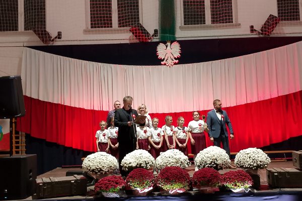 Zespół śpiewający na scenie z biało-czerwoną dekoracją i kwiatami podczas Festiwalu Pieśni Patriotycznych.