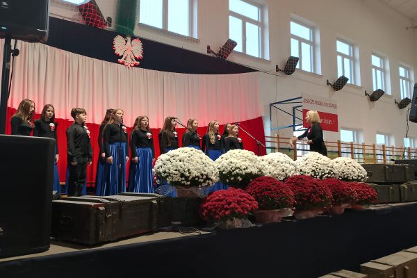 Zespół śpiewający na scenie z biało-czerwoną dekoracją i kwiatami podczas Festiwalu Pieśni Patriotycznych.