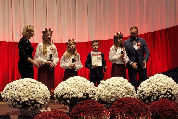 Zespół śpiewający na scenie z biało-czerwoną dekoracją i kwiatami podczas Festiwalu Pieśni Patriotycznych.