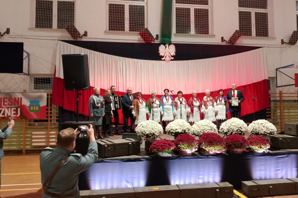 Zespół śpiewający na scenie z biało-czerwoną dekoracją i kwiatami podczas Festiwalu Pieśni Patriotycznych.