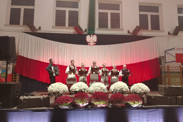 Zespół śpiewający na scenie z biało-czerwoną dekoracją i kwiatami podczas Festiwalu Pieśni Patriotycznych.