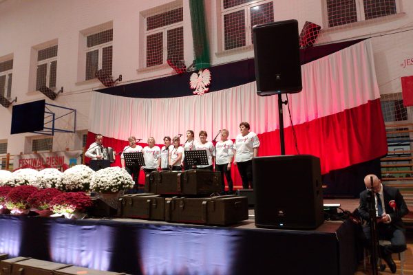 Zespół śpiewający na scenie z biało-czerwoną dekoracją i kwiatami podczas Festiwalu Pieśni Patriotycznych.