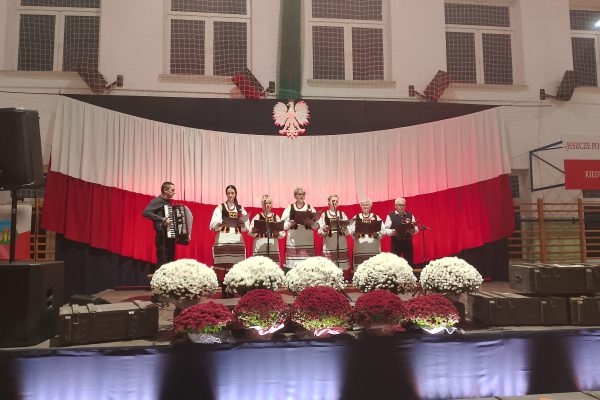 Zespół śpiewający na scenie z biało-czerwoną dekoracją i kwiatami podczas Festiwalu Pieśni Patriotycznych.