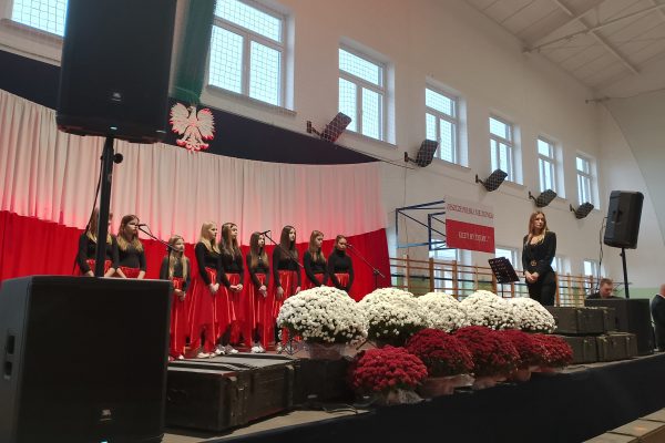 Zespół śpiewający na scenie z biało-czerwoną dekoracją i kwiatami podczas Festiwalu Pieśni Patriotycznych.