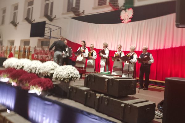 Zespół śpiewający na scenie z biało-czerwoną dekoracją i kwiatami podczas Festiwalu Pieśni Patriotycznych.