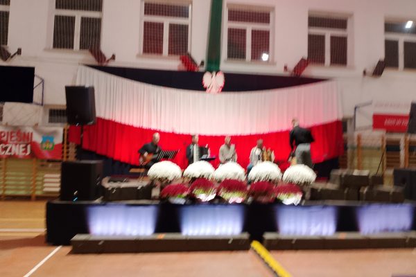 Zespół śpiewający na scenie z biało-czerwoną dekoracją i kwiatami podczas Festiwalu Pieśni Patriotycznych.