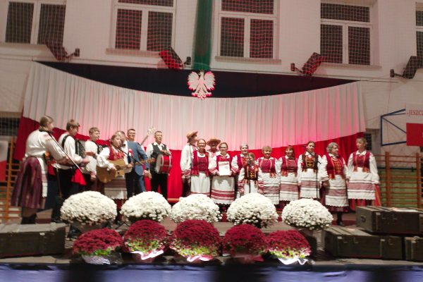 Zespół śpiewający na scenie z biało-czerwoną dekoracją i kwiatami podczas Festiwalu Pieśni Patriotycznych.