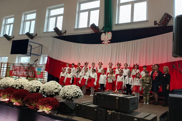 Zespół śpiewający na scenie z biało-czerwoną dekoracją i kwiatami podczas Festiwalu Pieśni Patriotycznych.