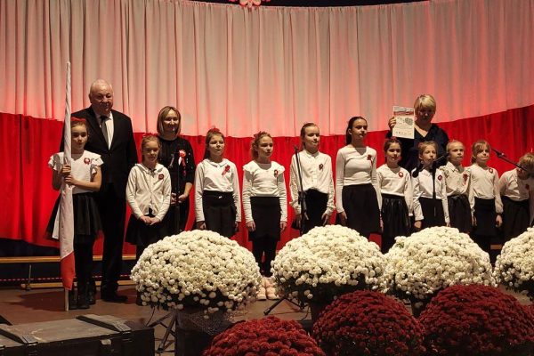 Zespół śpiewający na scenie z biało-czerwoną dekoracją i kwiatami podczas Festiwalu Pieśni Patriotycznych.